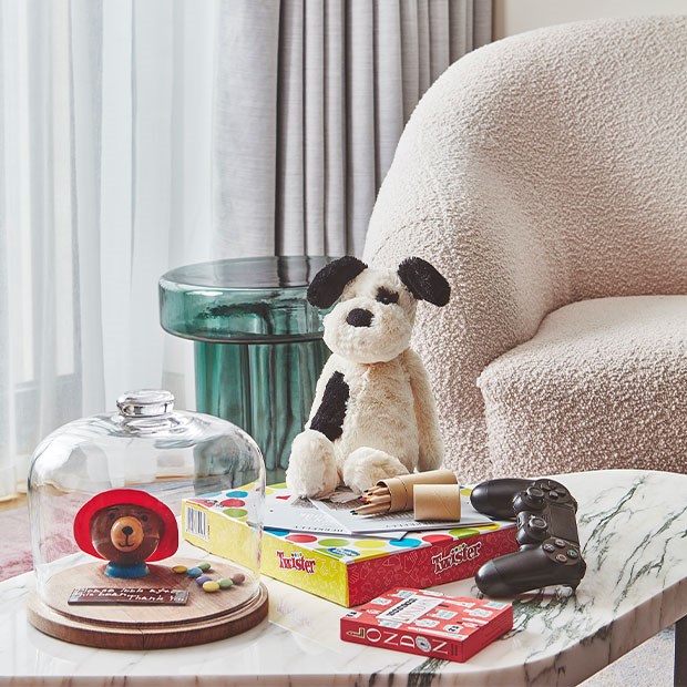 Jellycat toy dog, paddington bear cake, twister, colouring pencils, London game and games console controller on table with chair behind in suite at The Berkeley.