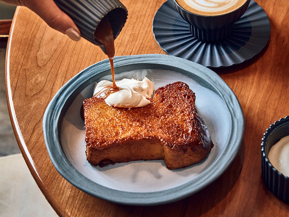 French toast with syrup being poured over. A small side dish of cream and a flat white are also fatured.