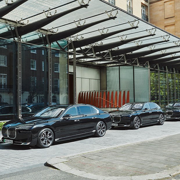 Side view of three black luxury sedans lined up outside a hotel entrance, reflecting surrounding buildings in the glass facade behind them.