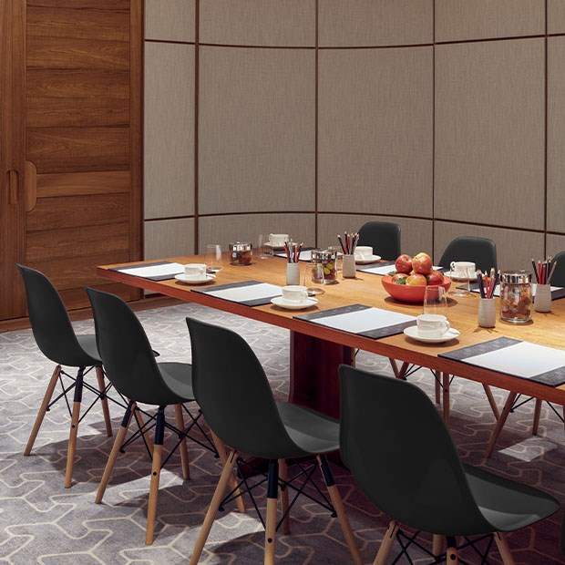 A section of a long conference table set up for a meeting, with neatly arrange pencils and notepads and bowls of snacks including fruit. There are black chairs tucked behind the table, wooden walls and light patterned carpet.
