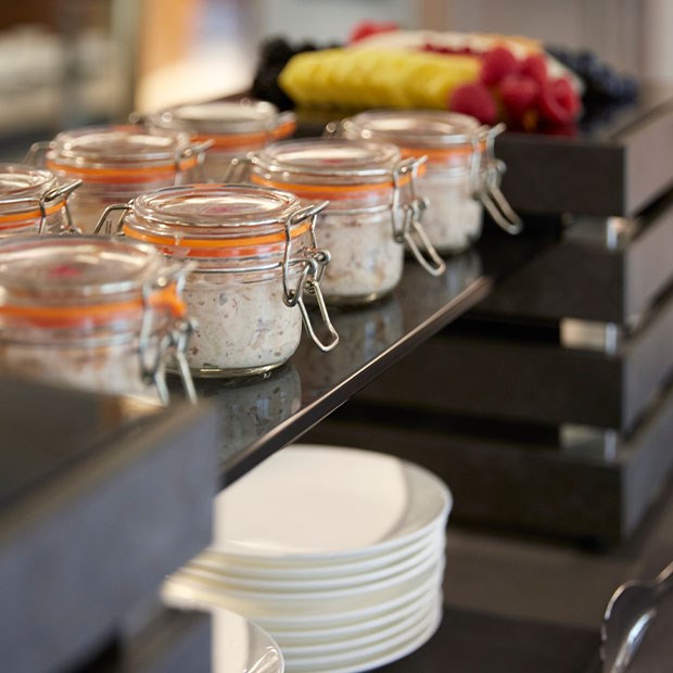 Image of a section of a breakfast spread, with mini jars of overnight oats in focus, and on the shelf below are a stack of white plates. Out of focus in the background is a big platter of chopped fruit.