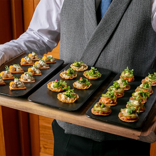Outline of man in grey waistcoat and white gloves holding a grey slate with a platter of canapes.