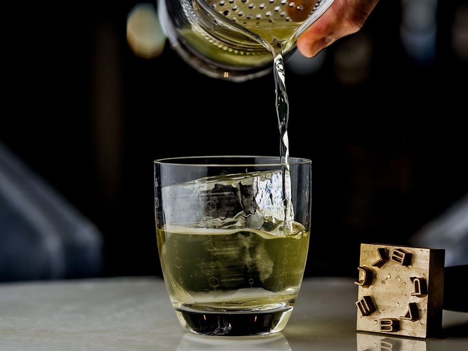 Green-coloured cocktail being poured through a strainer into a short glass with a Blue Bar ice stamp sat beside