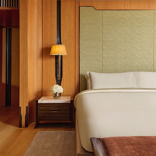 Tranquil bedroom corner with a soft neutral-toned bed, pale green fabric headboard, bedside table with fresh white flowers, and warm wood panelled walls.