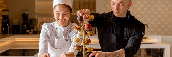 Two smiling chefs at The Berkeley carefully plate an artistic afternoon tea stand with intricate pastries, inside a bright, modern patisserie kitchen.