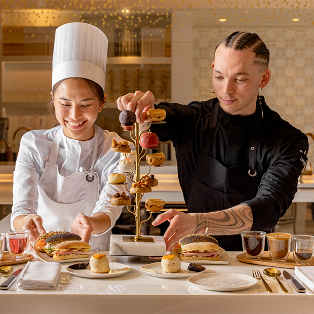Two smiling chefs at The Berkeley carefully plate an artistic afternoon tea stand with intricate pastries, inside a bright, modern patisserie kitchen.