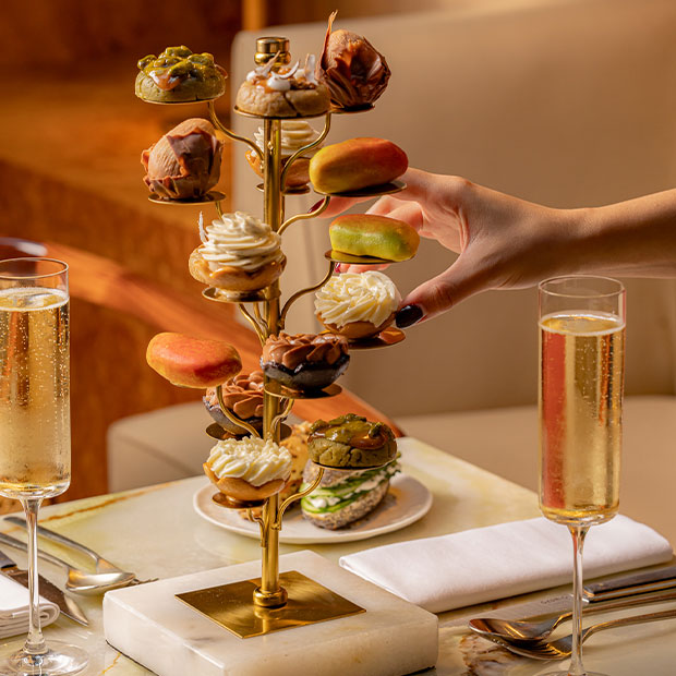 Hand selecting a miniature pastry from a tiered golden stand, with champagne glasses beside it.