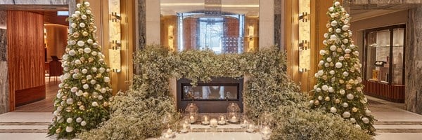 Festive lobby with two decorated Christmas trees, white florals around a fireplace, and candlelight.