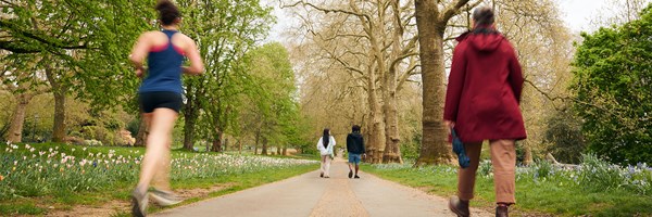 View of path in Hyde Park surrounded by trees and flowers. Runners and passers by can be seen making their way through the park.