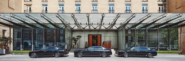 Three black luxury cars parked in front of a modern glass canopy entrance, with a grand hotel facade and symmetrical windows above.