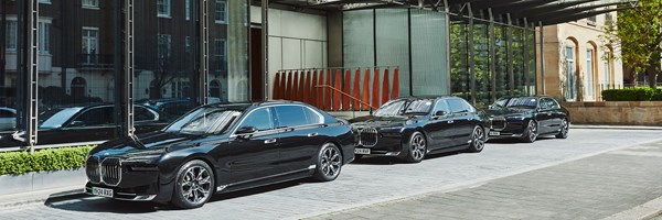 Side view of three black luxury sedans lined up outside a hotel entrance, reflecting surrounding buildings in the glass facade behind them.