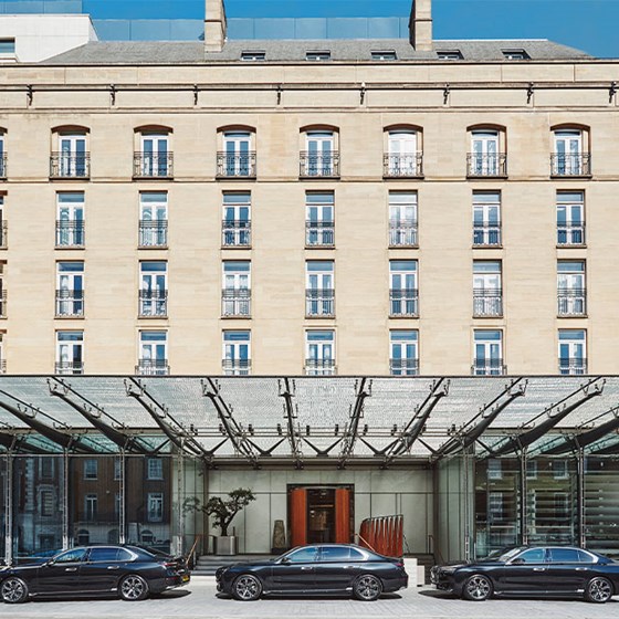 Three black luxury cars parked in front of a modern glass canopy entrance, with a grand hotel facade and symmetrical windows above.