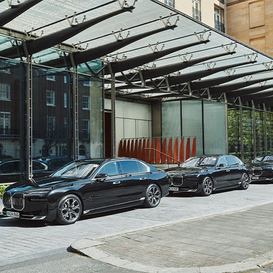 Side view of three black luxury sedans lined up outside a hotel entrance, reflecting surrounding buildings in the glass facade behind them.