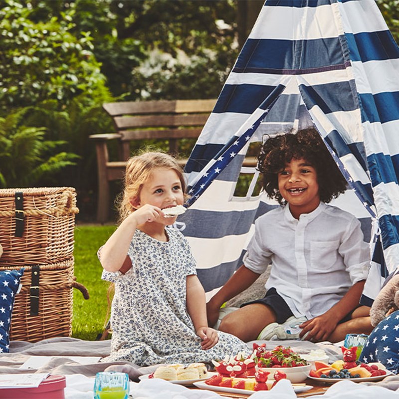 A little girl and boy, having a picnic on the grass, with a striped teepee and traditional picnic baskets behind them. The scene is summery.