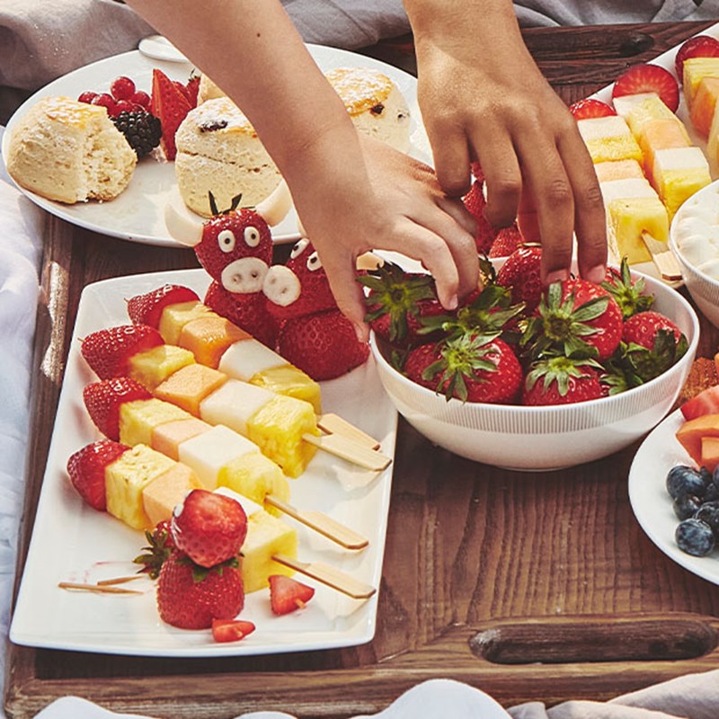 A Berkeley children's pincic, with two little hands reaching into a bowl of strawberries. There are also fruit kebabs and scones.