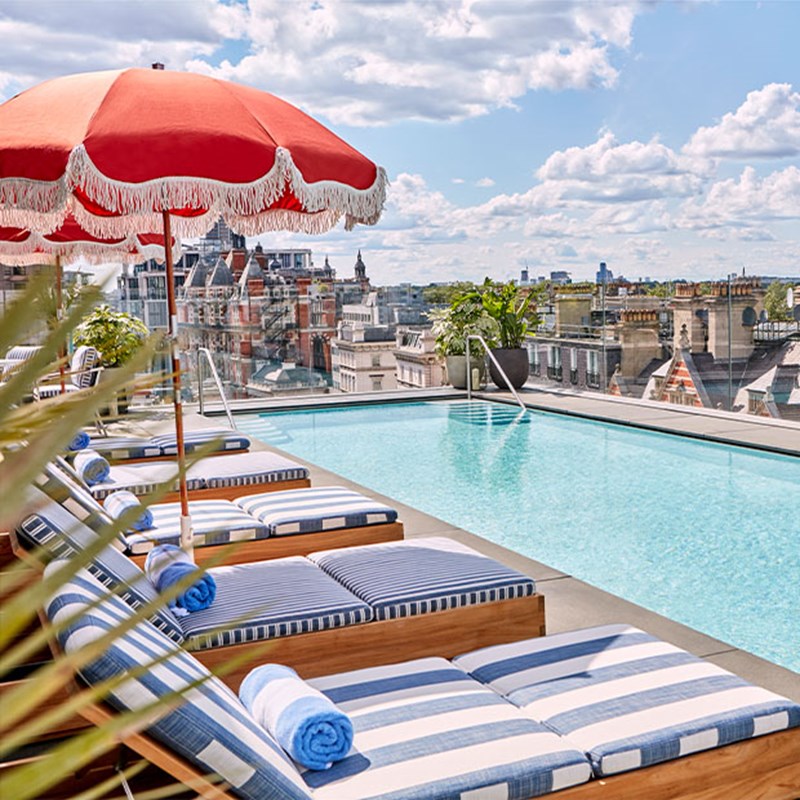The Berkeley Rooftop Pool, with blue & white striped sun loungers and red parasols. A view of London's urban landscape in the background.