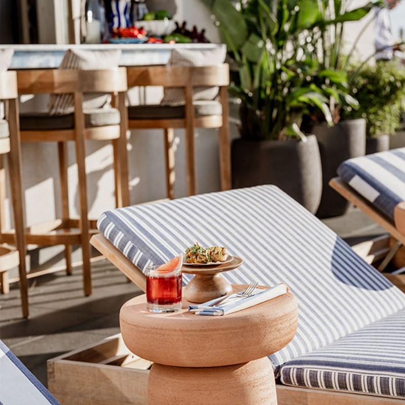 Small table next to a blue & white striped sun lounger at The Berkeley Rooftop Bar, with a napkin, knife & fork, a negroni and a small plate with a seafood snack.