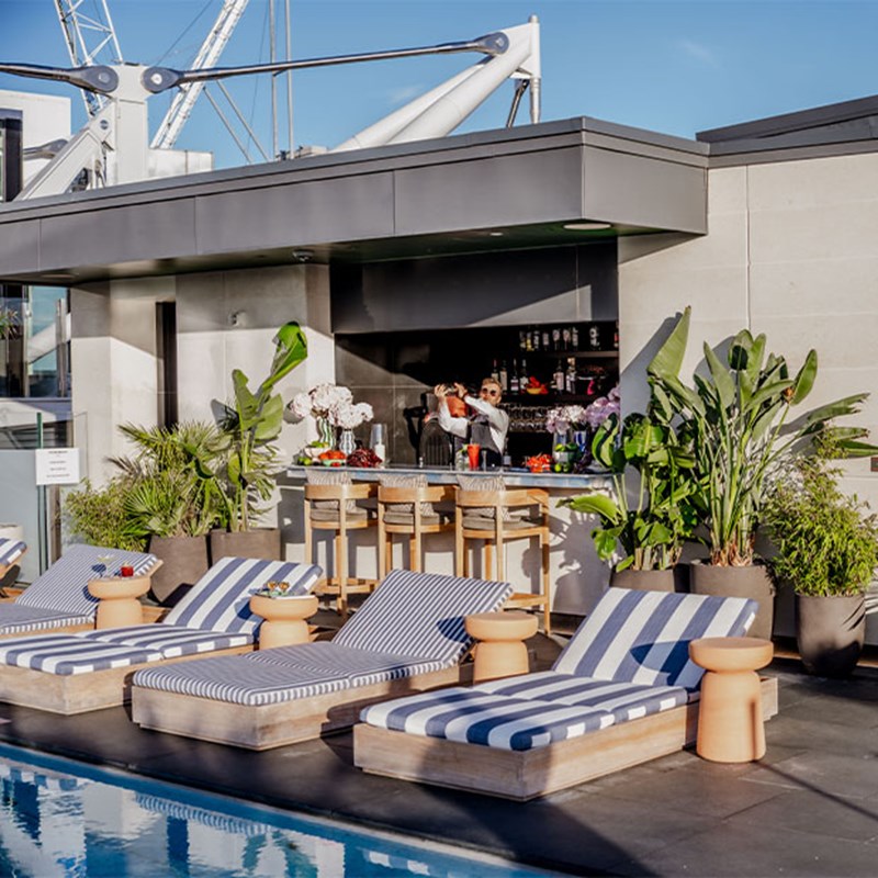 Side on view of The Berkeley Rooftop Bar, with a bit of the pool in view, and blue & white striped sunloungers, with the bar behind and a barman shaking a cocktail.