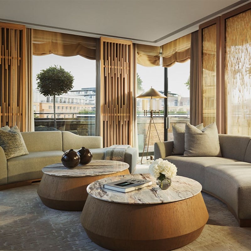 A sophisticated lounge with curved sofas, marble-topped wooden tables, and floor-to-ceiling windows overlooking a cityscape with greenery outside.