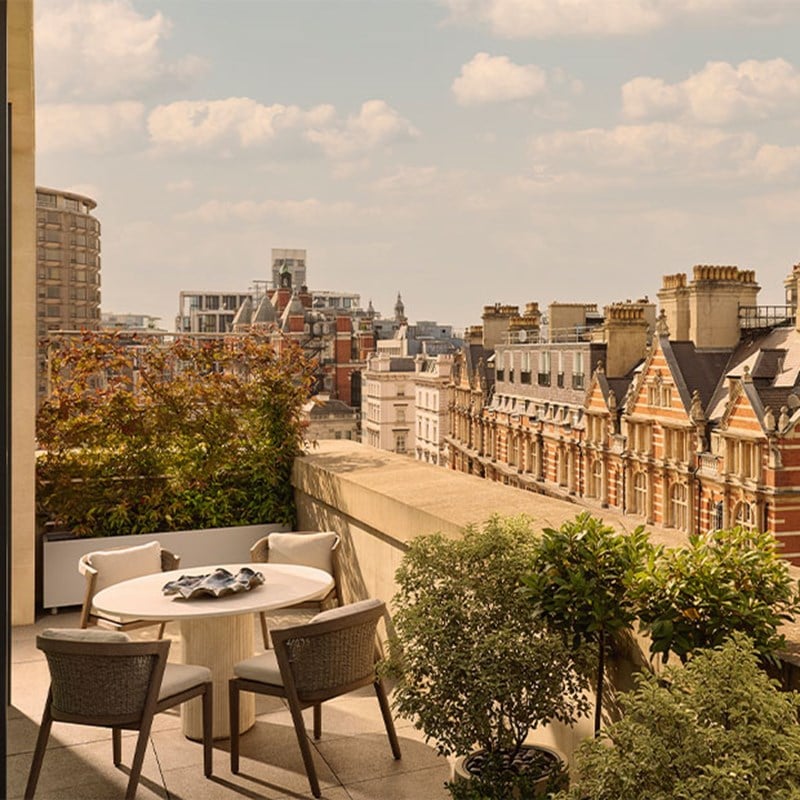 An outdoor terrace with a round dining table, chairs, lush green plants, and views of historic Knightsbridge rooftops under a blue sky.
