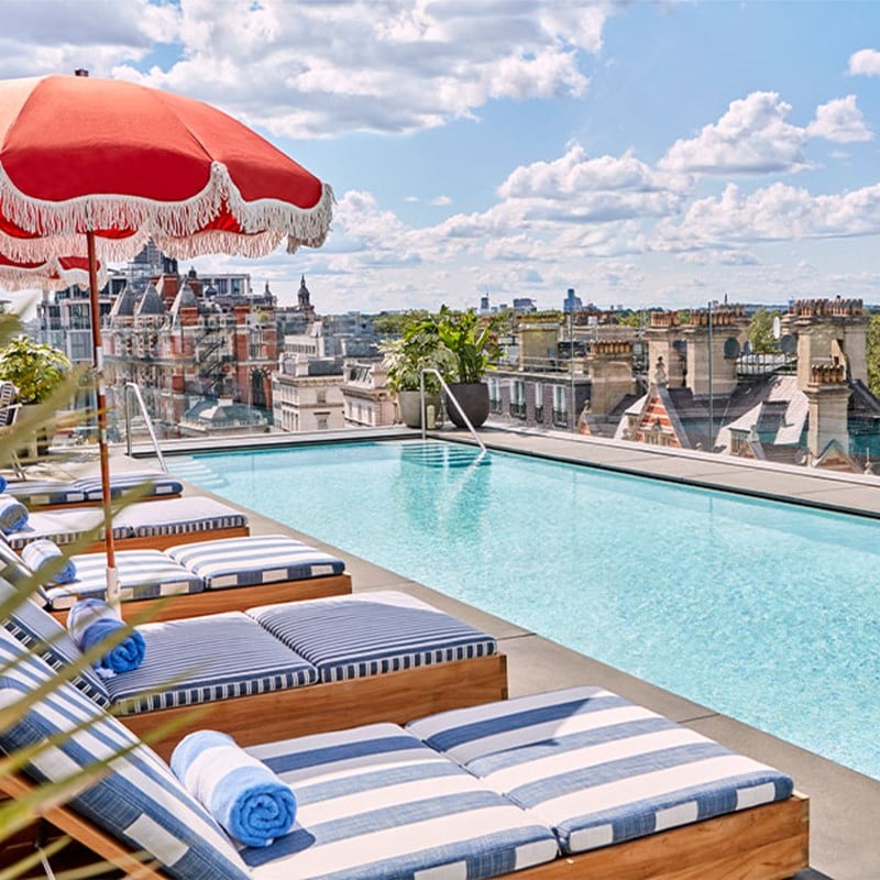 A rooftop swimming pool lined with striped loungers, a red parasol, and sweeping views of London's iconic historic rooftops.