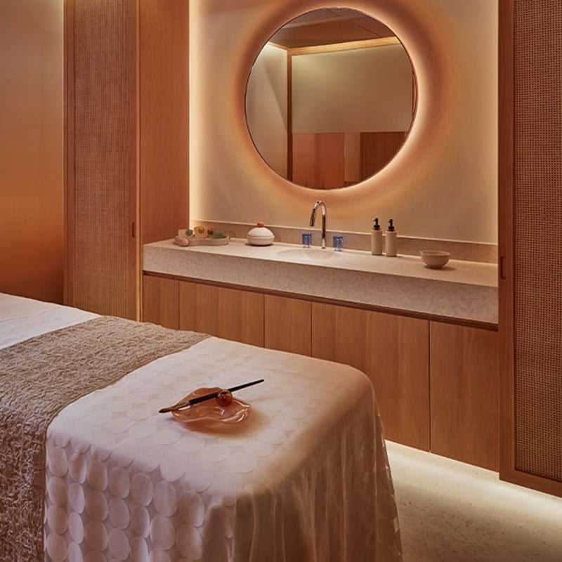 A serene spa treatment room with soft lighting, a massage bed draped in white linen, and a minimalist vanity with a round mirror.