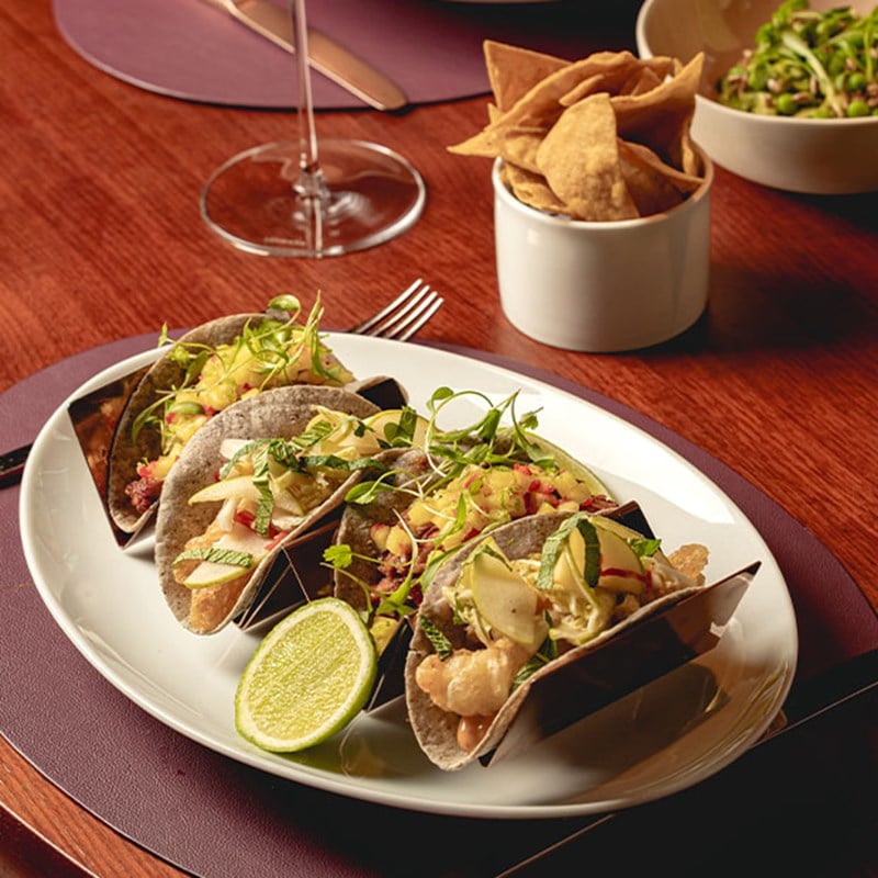 A plate of gourmet tacos with fresh herbs and lime, accompanied by a bowl of tortilla chips on a polished wooden table