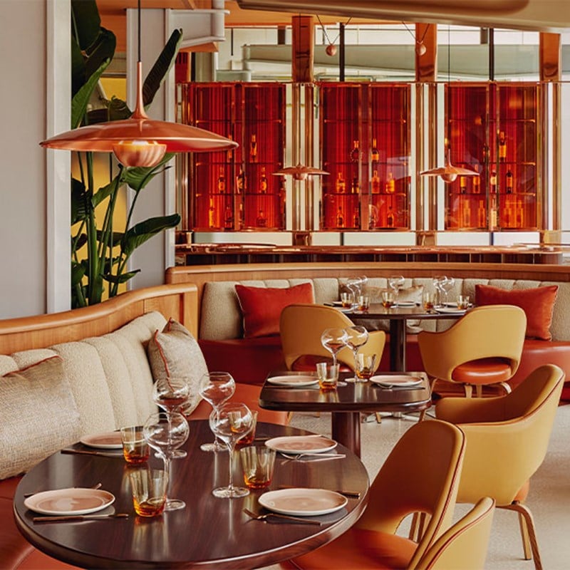 A lively restaurant interior with soft curved seating, amber-toned accents, and floor-to-ceiling glass shelving displaying bottles behind the tables.