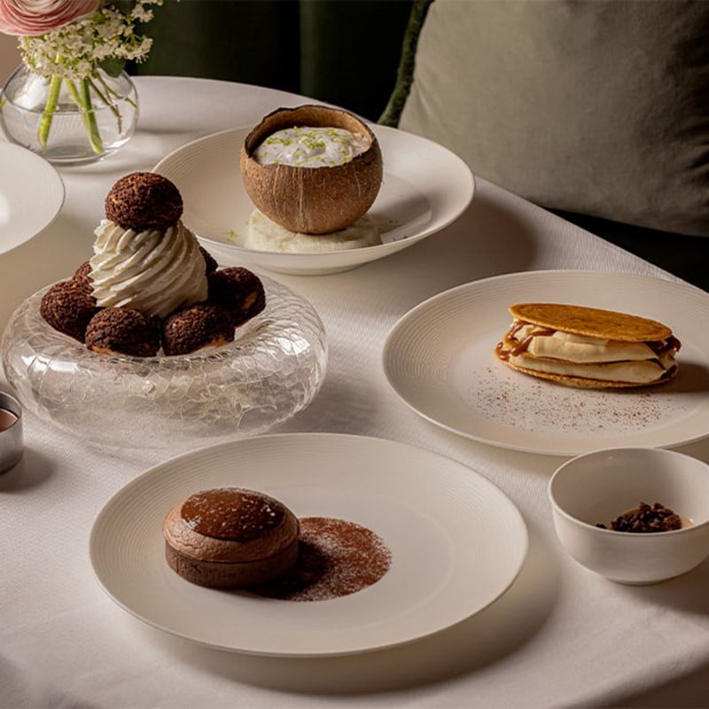 An elegant dessert table with plates of intricate pastries, chocolate fondant, coconut mousse, and fresh flowers on a crisp white tablecloth.