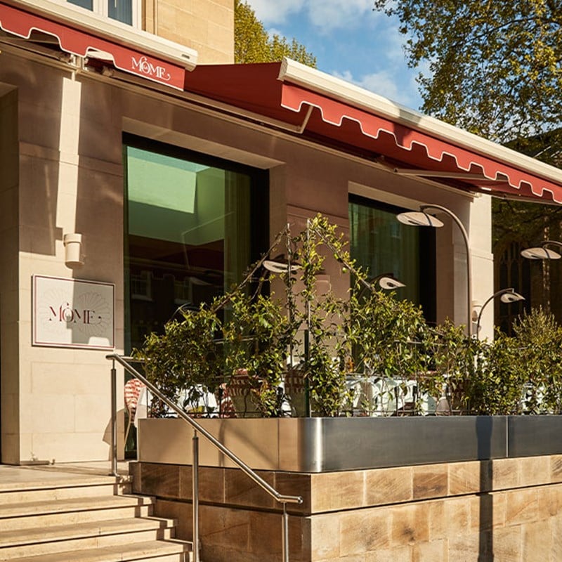 Street-level view of the La Môme restaurant exterior with red awnings, large windows, and a planted terrace along the entrance steps.