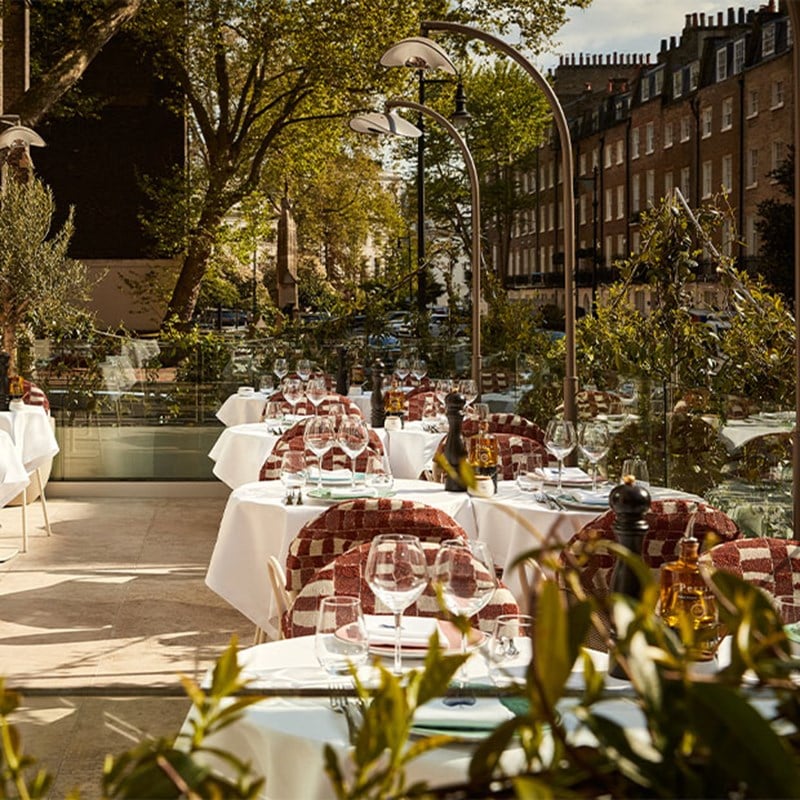 Elegant outdoor dining terrace with white tablecloth-covered tables, red-patterned chairs, and street views framed by greenery and curved lamps.
