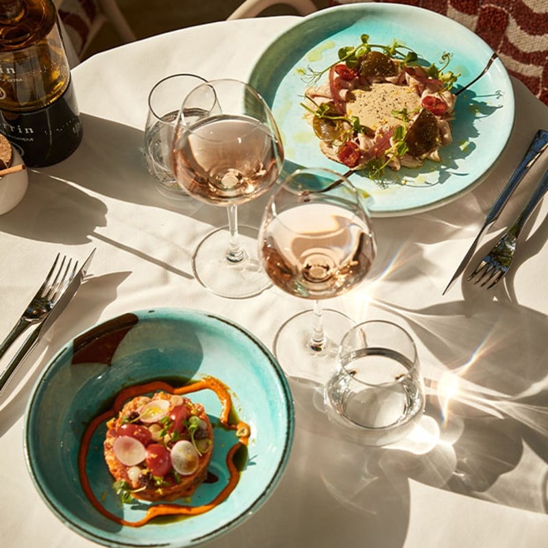Overhead view of a restaurant table with two vibrant plated dishes, three glasses of rosé wine, cutlery, and sunlight casting soft shadows.