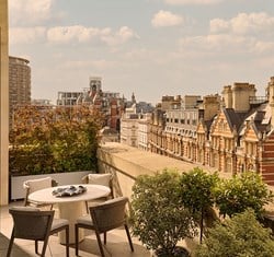 Rooftop dining area with a round table and four chairs overlooking classic London rooftops and architectural details under a bright sky.