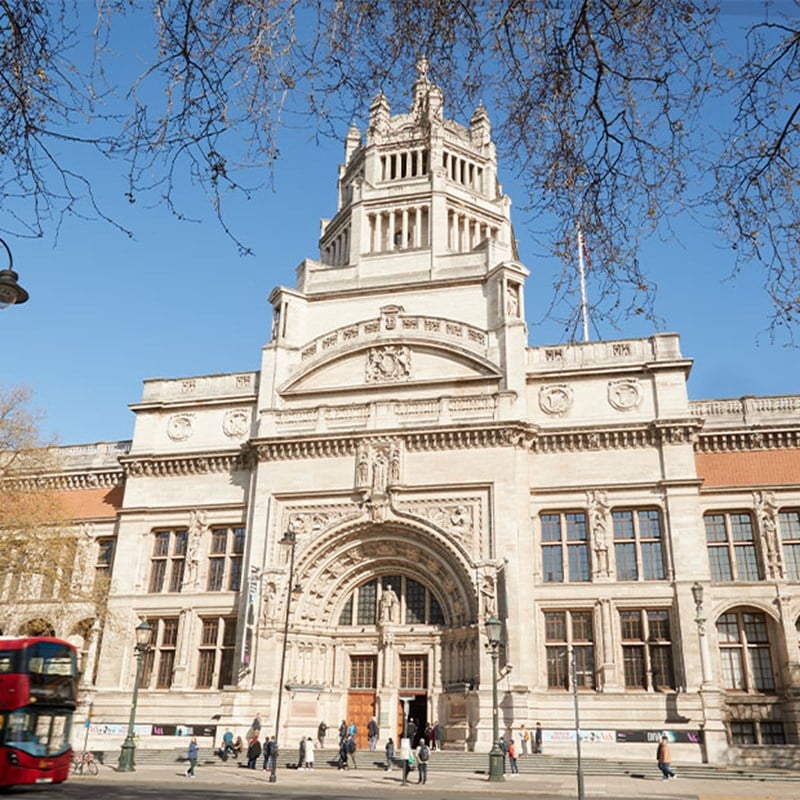 The grand entrance of the Victoria and Albert Museum in London, showcasing intricate stonework, a large arched doorway, and visitors outside.