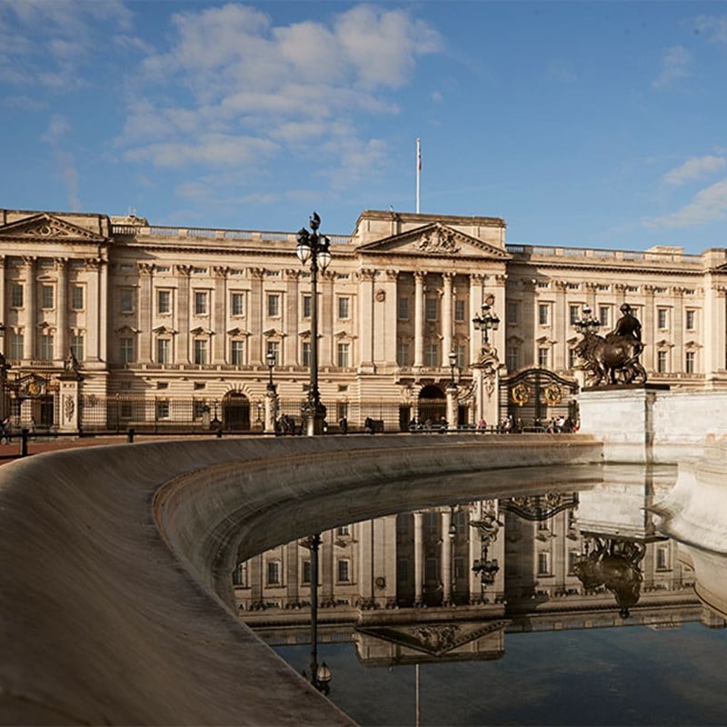 View of Buckingham Palace and the Victoria Memorial reflected in the curved fountain basin on a sunny day.