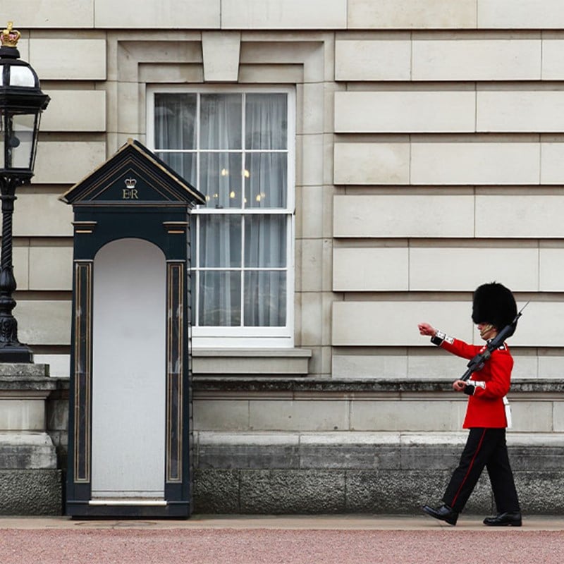 A Queen’s Guard in red uniform and bearskin hat marches beside a black sentry box outside Buckingham Palace.