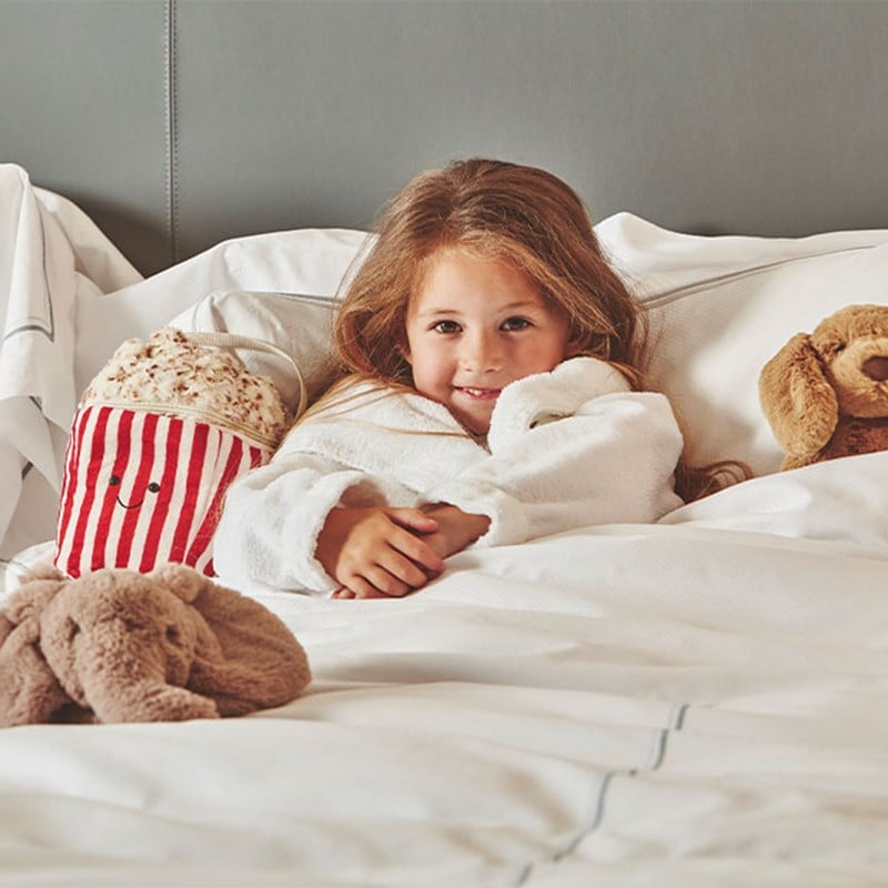 A young child wrapped in a fluffy robe, nestled in a bed surrounded by plush toys, with a warm smile that conveys the comfort and joy of the holiday season.