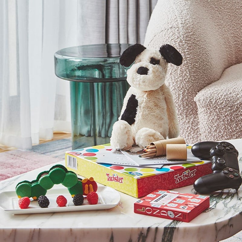 A cozy setup featuring a plush dog toy, board games, a gaming controller, and a plate of colorful treats, set on a marble table with a soft, inviting backdrop.