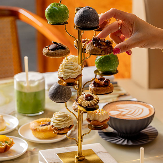 A luxurious golden dessert tree, presenting an assortment of pastries adorned with cream swirls, chocolate garnishes, and whole green and black fruits, with a hand delicately reaching for one treat.