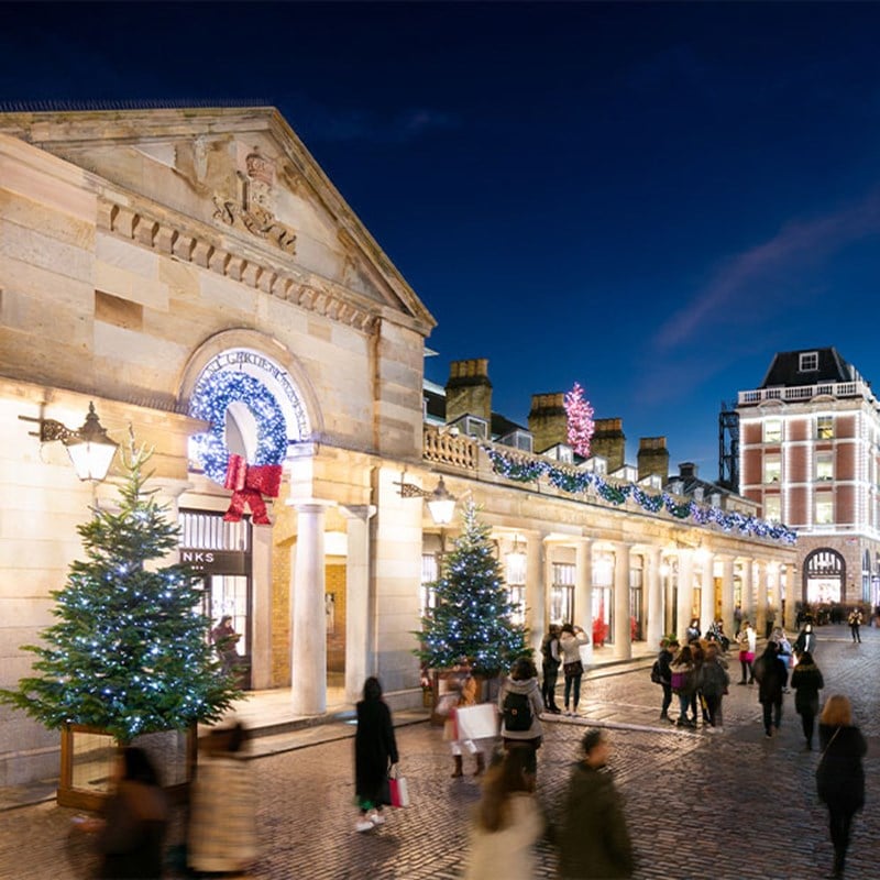 A festive evening scene at Covent Garden, adorned with Christmas lights and decorations, with people enjoying the atmosphere.