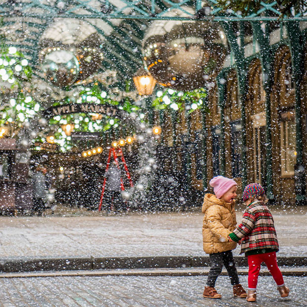 Two young children dressed in winter attire delight in the falling snow at Covent Garden, surrounded by holiday decorations.