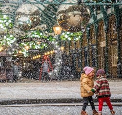 Two young children dressed in winter attire delight in the falling snow at Covent Garden, surrounded by holiday decorations.