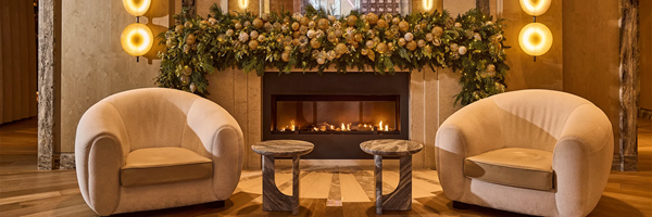 Festive hotel lounge with armchairs facing a fireplace, framed by garlands, gold baubles, and warm lighting.