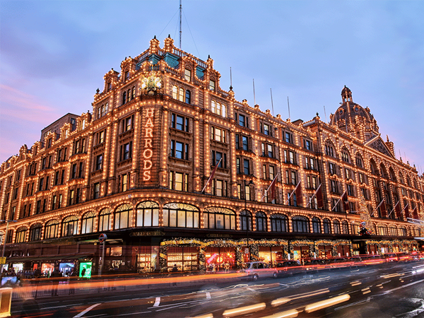 Harrods department store illuminated with festive lights at dusk, overlooking a busy London street.