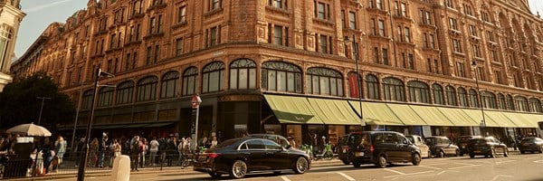 Exterior view of Harrods department store with people walking on the pavement and cars driving past.