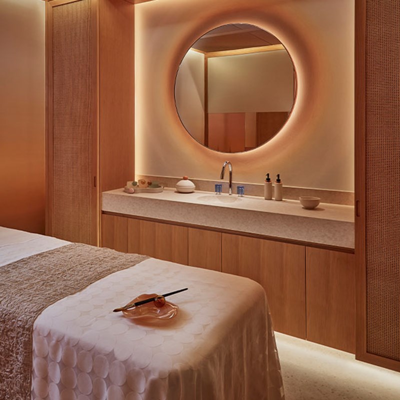 Calming spa treatment room with a massage bed, wooden cabinets, and a round illuminated mirror.