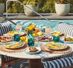 Vibrant poolside dining table set with colourful plates, yellow napkins, floral centrepiece, and Mediterranean-style dishes.
