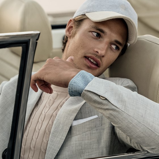 A man wearing a white knitwear, a jacket and a cap in a car.