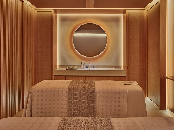 A serene spa treatment room with two massage tables, soft lighting, and a circular mirror above a minimalist vanity.