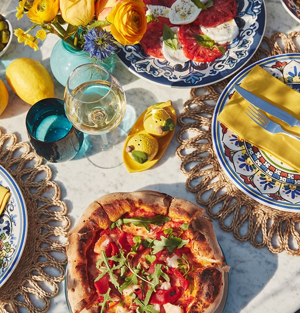 Vibrant Italian-style summer table setting at The Berkeley’s rooftop, featuring a wood-fired pizza topped with rocket and peppers, a caprese salad of tomatoes, mozzarella, and basil on a decorative blue plate, and a glass of white wine. The table is adorned with colourful ceramic plates, bright yellow napkins, lemons, a floral arrangement, and playful lemon-shaped salt and pepper shakers, evoking a sunny Capri-inspired atmosphere.
